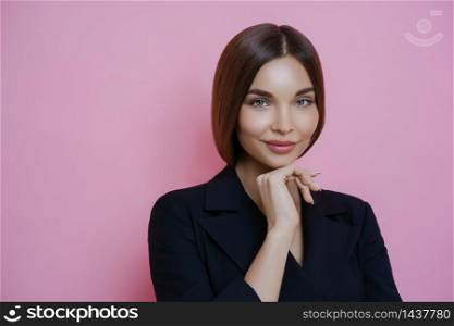 Photo of attractive well groomed businesswoman wears elegant black costume, keeps hand under chin, looks calmly at camera, has gentle look, going to have business meeting isolated over rosy background
