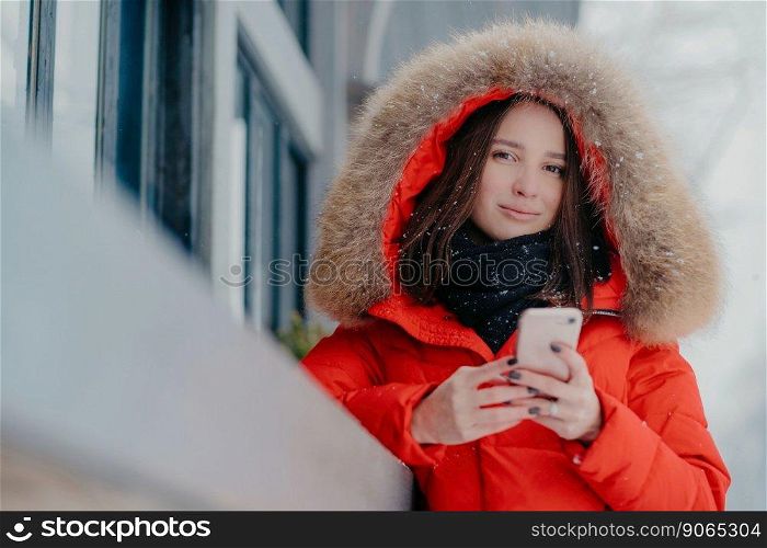 Photo of attractive woman holds modern modern gadget, wears red jacket with hoody, checks email box, connected to wireless internet, stands outdoor, enjoys free time and frosty winter weather