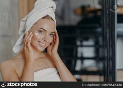 Photo of attractive woman touches face and applies face cream has natural makeup and manicure wears bath towel wrapped on head has healthy pure skin, poses indoor. Female model with fresh daily makeup