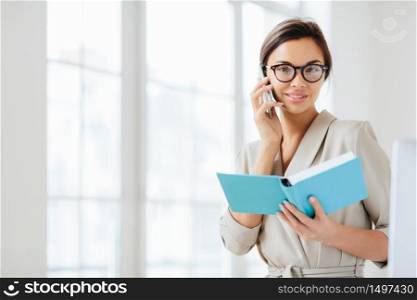 Photo of attractive woman with dark hair, has telephone conversation, agrees to meet with colleague, holds opened diary, wears glasses and formal suit, poses over white background plans daily schedule