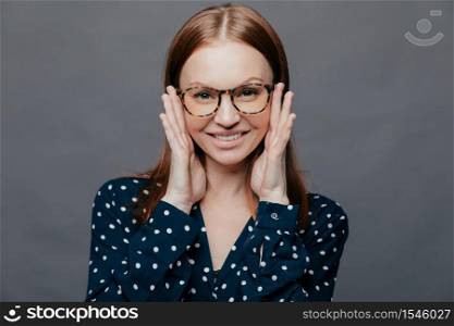 Photo of attractive woman with pleased facial expression, keeps both hands near cheeks, wears polka dot shirt, poses over grey studio wall, being happy. European pleased lady expresses happiness