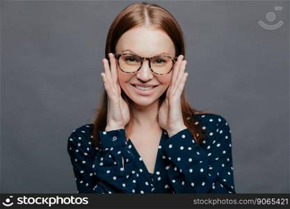 Photo of attractive woman with pleased facial expression, keeps both hands near cheeks, wears polka dot shirt, poses over grey studio wall, being happy. European pleased lady expresses happiness