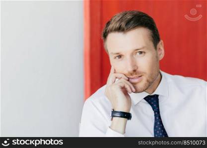 Photo of attractive young businessman with confident expression holds chin, dressed in formal white shirt, has watch on arm, thinks about starting new business, poses against red and white wall