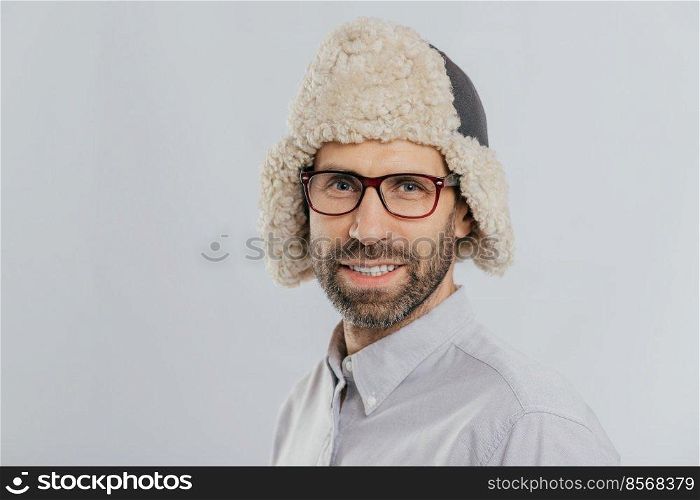 Photo of attractive young European male with pleasant appearance, wears warm fut hat, transparent glasses, models over white studio wall, rejoices shopping during weekend. People, style concept
