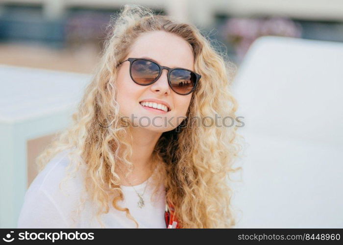 Photo of attractive young female has light curly hair, wears trendy shades, has toothy smile, shows white teeth, recreats outdoor, smiles as recieves compliment from boyfriend. Summer time, rest
