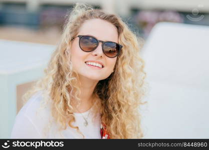 Photo of attractive young female has light curly hair, wears trendy shades, has toothy smile, shows white teeth, recreats outdoor, smiles as recieves compliment from boyfriend. Summer time, rest