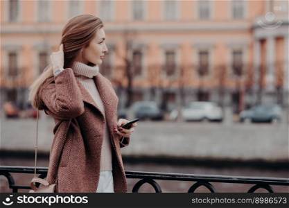 Photo of attractive young female model with long hair, dressed in ncoat, has outdoor stroll during autumn time in big city, holds modern cell phone, text messages, admires beautiful cityscape