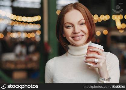 Photo of attractive young woman holds takeaway coffee, has pleased expression, toothy smile, dressed in white jumper, poses outside with blank space for your promotional content or advertisement