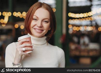 Photo of attractive young woman holds takeaway coffee, has pleased expression, toothy smile, dressed in white jumper, poses outside with blank space for your promotional content or advertisement