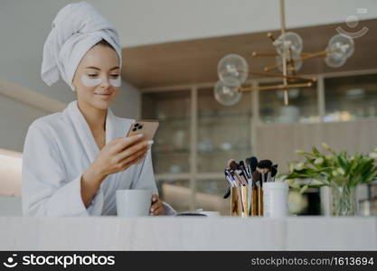 Photo of attractive young woman wears bath towel on head and soft white dressing gown holds mobile phone enjoys online communication applies moisturising undereye patches drinks hot beverage