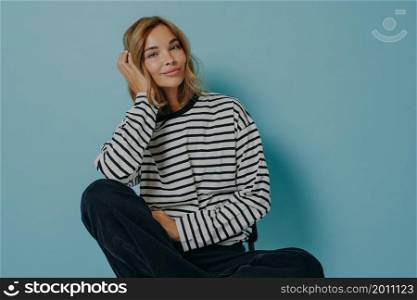 Photo of attractive young woman with minimal makeup sits relaxed on chair wears casual striped jumper and black trousers leans head on hand looks at camera satisfied isolated over blue background. Photo of attractive young woman with minimal makeup sits relaxed on chair over blue background