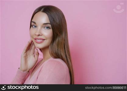 Photo of beautiful brown haired Caucasian woman keeps hand on cheek, smiles gently and looks directly at camera, has blue eyes, wears minimal makeup, stands in profile against pink background