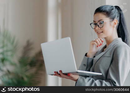 Photo of beautiful brunette woman holds chin, concentrated into laptop screen, studies financial market, focused on remote job, prepares publication. Freelacer watches webinar to improve skills