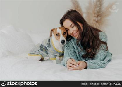 Photo of beautiful brunette woman in knitted sweater lies together wih dog on soft bed, enjoys spending time with favorite pet, cares about animals, stay at home during coronavirus quarantine