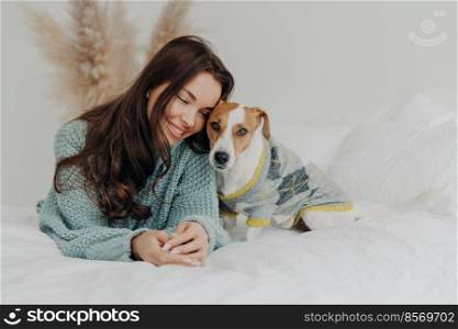 Photo of beautiful brunette woman in knitted sweater lies together wih dog on soft bed, enjoys spending time with favorite pet, cares about animals, stay at home during coronavirus quarantine