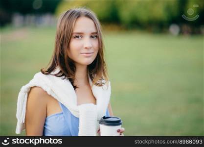 Photo of beautiful comely girlfriend with white sweater on shoulders, drinks takeaway coffee, thinks about something, stands outdoor, has walk across green field. People, lifestyle and spare time
