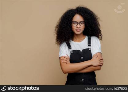 Photo of beautiful dark skinned teenage girl poses with arms crossed, wears spectacles, white t shirt and black sarafan, stands against brown background, copy space for your promotional content