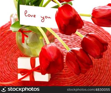Photo of beautiful red tulip flowers bouquet and romantic postcard in it with small white gift box on the table, springtime holiday, loving message, present for birthday, happy mothers day&#xA;