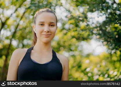 Photo of beautiful sporty woman in sportswear uses wireless earphone for listening music rests in green park looks directly at camera has rest after outdoor training involved in sport activities