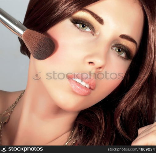 Photo of beautiful stylish woman doing makeup, closeup portrait of pretty female with silky brown hair holding brush for blush isolated on grey background, smoky eyes, fashionable makeover