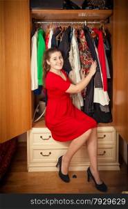 Photo of beautiful woman in red dress sitting at big wardrobe