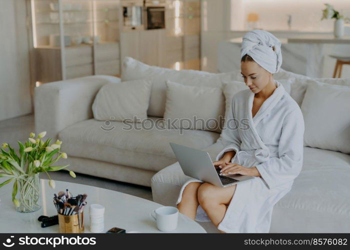 Photo of beautiful young woman in bathrobe types on laptop computer works distantly from home poses on comfortable couch against modern apartment interior drinks tea. People wellbeing lifestyle