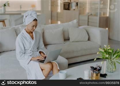 Photo of beautiful young woman in bathrobe types on laptop computer works distantly from home poses on comfortable couch against modern apartment interior drinks tea. People wellbeing lifestyle