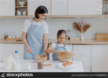 Photo of brunette mother and little kid make cookie dough, whisk ingredients in bowl, dressed in aprons, mum rejoices having small helper at kitchen. Cooking together, family and home concept