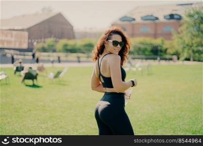 Photo of brunette sporty woman holds bottle of water dressed in active wear has rest after training smiles pleasantly poses outdoor against green lawn. Morning workout and healthy lifestyle concept