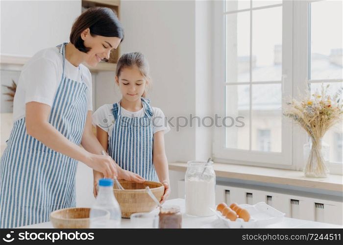 Photo of brunette woman in striped apron, mixes ingredients with beater, shows small daughter how to cook, stand at kitchen near table with fresh products. Mother and child busy preparing meal