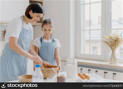 Photo of brunette woman in striped apron, mixes ingredients with beater, shows small daughter how to cook, stand at kitchen near table with fresh products. Mother and child busy preparing meal