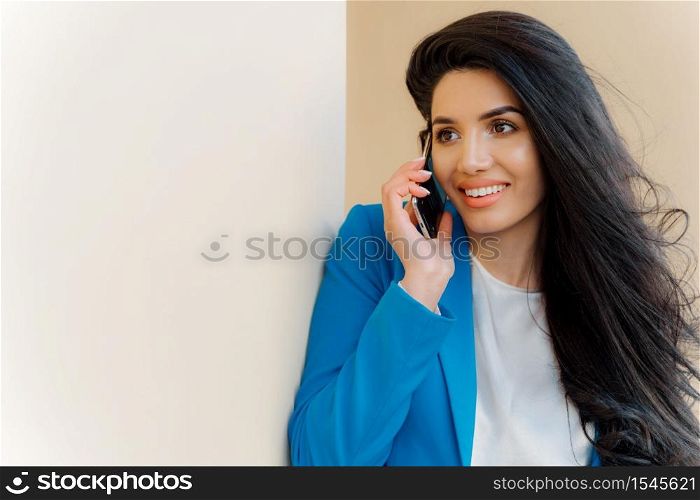 Photo of brunette woman with pleasant appearance, luxurious hair, talks on mobile phone, dressed in formal clothes, arranges business meeting with partner. People, technology, communication concept