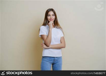 Photo of brunette young European woman holds chin, wears white t shirt and jeans, enjoys conversation with her mates, isolated over brown background. People, lifestyle, face expressions concept