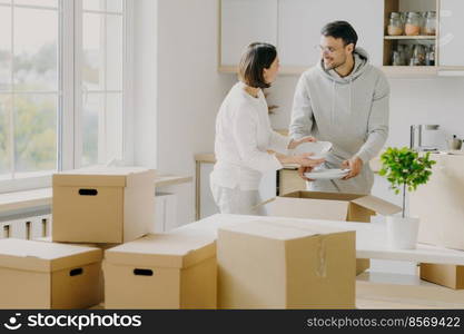 Photo of busy family couple unpack personal stuff from carton boxes, dressed in casual clothes, hold white plates, pose in spacious kitchen with modern furniture, surrounded with pile of packages