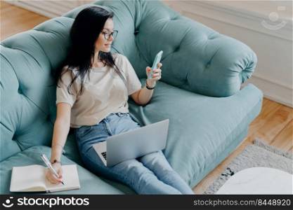 Photo of busy female freelancer in casual clothes focused in smartphone display, checks information in internet, notes something in diary, uses modern technologies for work, poses at cozy room