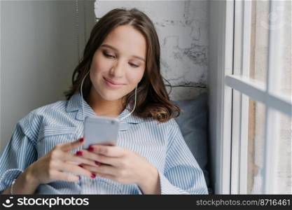 Photo of Cauasian woman selects favourite song in playlist, uses contemporary cellular and high quality earphones, dressed in casual outfit, poses on window sill in cozy room alone. Technology