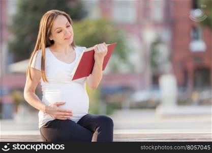 Photo of caucasian brunette pregnant woman reading the book on a bench