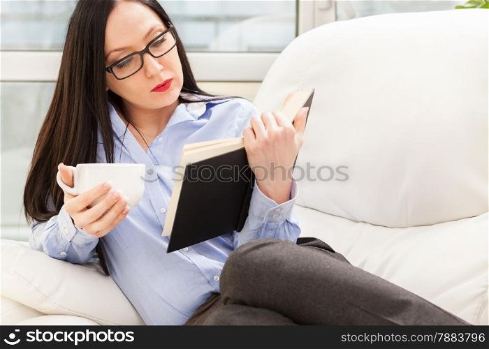 Photo of caucasian businesswoman reading a book while relaxing on the sofa with cup of coffee