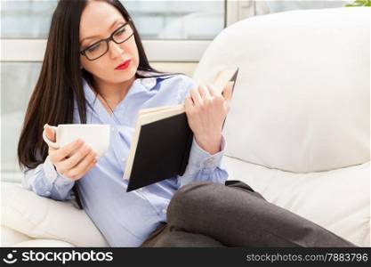 Photo of caucasian businesswoman reading a book while relaxing on the sofa with cup of coffee