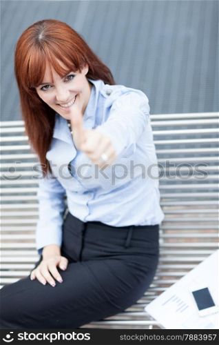 Photo of caucasian businesswoman sitting on a metal bench and making positive thumb gesture