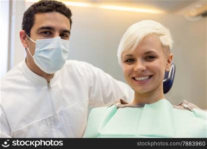photo of caucasian male dentist sitting next to his smiling female patient and looking towards the camera
