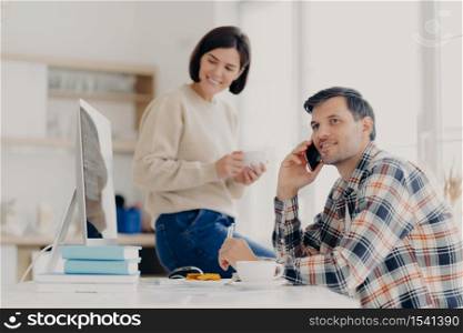 Photo of cheerful busy male freelancer uses mobile phone, discusses latest news, sits at desktop with papers and modern computer. Positive woman dressed in sweater and jeans, holds mug of drink
