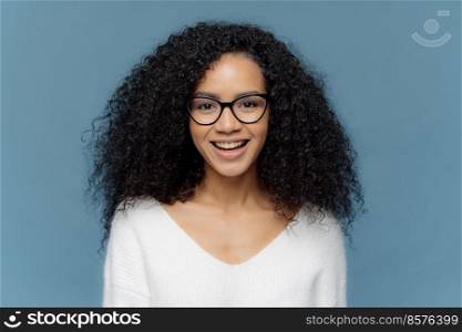 Photo of cheerful dark skinned female with bushy crisp hair, smiles happily at camera, rejoices nice day, dressed in white sweater, isolated over blue background expresses positiveness. Human feelings