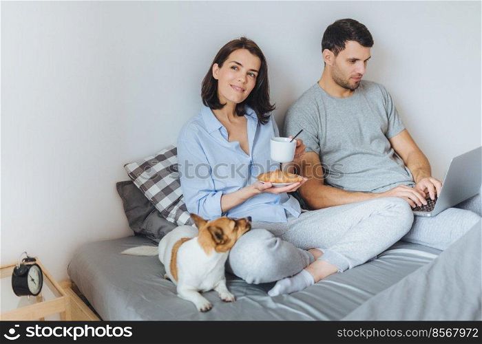 Photo of cheerful female drinks coffee with croissant, sits crossed legs with dreamful expression, has breakfast in bed and her pet, husband sits near, keyboards on laptop computer