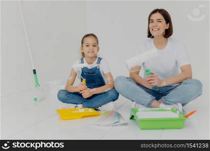 Photo of cheerful girl and daughter sit crossed legs on floor, hold paint rollers, paint room in white color, busy with refurbishment, have happy expressions. Home, repair, building and decoration