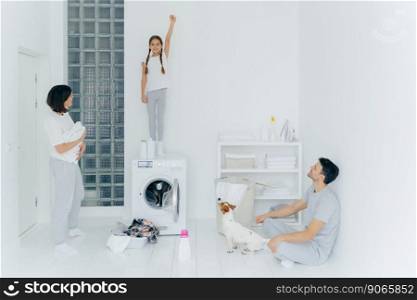 Photo of cheerful girl stands on washing machine, raises arm with clenched fist, helps parents to wash clothes, mother, father and dog look at kid. Family in laundry room, busy with domestic chores