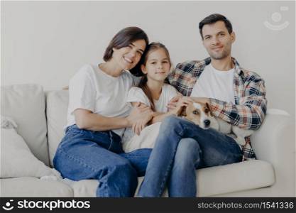 Photo of cheerful husband and wife spend free time with their lovely daughter and favourite domestic animal, sit all together at sofa, have good mood, enjoy togetherness, show love to each other