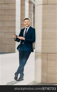 Photo of cheerful male dressed in expensive black suit, stands crossed legs, works on touch pad, stands in modern office interior, smiles positively at camera. Young male manager. Full length portrait