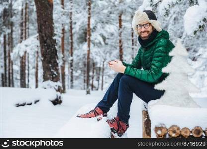 Photo of cheerful male types messages on smart phone looks directly into camera, sits in white winter forest, smiles happily, enjoys communication, watches video, admires landscapes