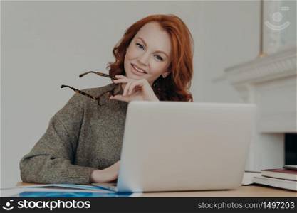 Photo of cheerful redhead female student makes research for homework, sits at coworking space in study room, works on laptop computer, uses internet. Freelance web designer creats design for client